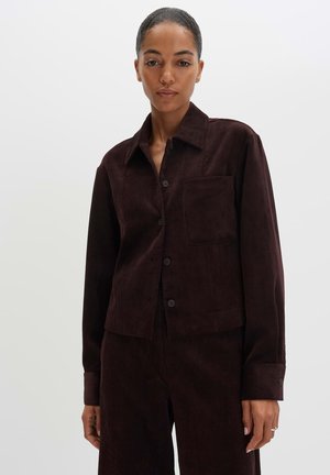 Vrouw met donker haar in een staart, gekleed in een donkerbruine ribfluwelen jas en bijpassende broek tegen een effen lichte achtergrond.
