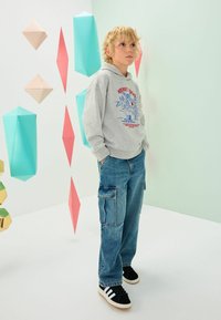 Grå hættebluse med trykt grafik, blå denim cargo-bukser og sorte sneakers med hvide striber. Geometrisk pastel dekoration i baggrunden.