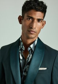 Chaqueta de esmoquin en teal con solapas de satén, llevada sobre una camisa de patrón en gris y blanco. Presenta un pañuelo blanco en el bolsillo, un cuello afilado y una tela texturizada.