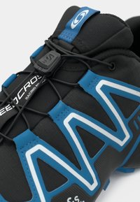 Μαύρο αθλητικό παπούτσι με μπλε λεπτομέρειες, υφασμάτινη επιφάνεια και σχέδιο ζιγκ-ζαγκ. Διαθέτει σύστημα στήριξης κορδονιών για σταθερή εφαρμογή. Ελαφρύς σχεδιασμός.