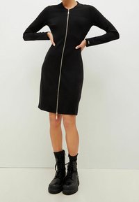 Svart långärmad klänning med en dragkedja framtill, figurnära design och rund halsringning. Matchad med knähöga svarta snörstövlar.