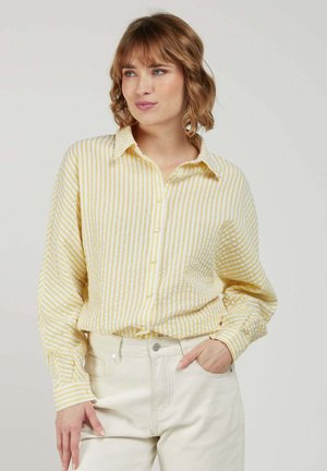 Femme aux cheveux courts et ondulés portant une chemise à boutons rayée jaune et blanche et un pantalon crème, une main dans la poche, regardant légèrement sur le côté.