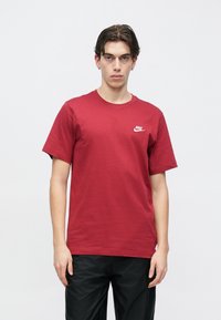 Mladi moški v rdeči majici Nike in črnih hlačah stoji ob belem ozadju, obrnjен naprej z nevtralnim izrazom na obrazu.