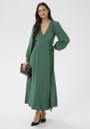 Kobieta stojąca w zielonej sukience kopertowej z długimi bufkami na rękawach, trzymająca czarną kopertówkę i nosząca czarne szpilki w szpic, uśmiechająca się do aparatu.