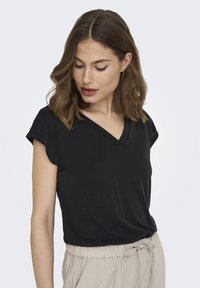 T-shirt noir à manches courtes et col en V, fabriqué en tissu lisse, porté avec un pantalon beige à cordon de serrage, présentant une coupe décontractée et un design simple.