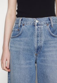 Personne portant un jean en denim bleu taille haute et un haut noir sans manches, la main détendue sur le côté.