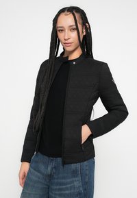 Mustast läbiõmmeldud bomberjope esil lukuga, püstitatud krae ja küljetaskutega, kombineeritud musta topi ja siniste teksadega.