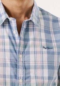 Detailní záběr na osobu v košili s kostkovaným vzorem, která má modrou, růžovou a bílou barvu a malou vyšitou nášivku na hrudi.