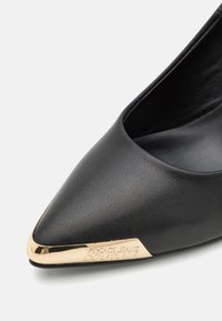 Chaussure en cuir noir à bout pointu avec un accent en métal doré au bout et un logo embossé. Texture lisse avec une silhouette élégante.