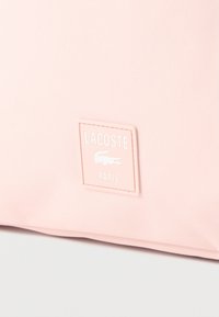 Svetloružová látka so štvorcovou štítkom zošitým na nej, s nápisom "LACOSTE", logom aligátora a nápisom "PARIS".