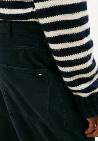 Le salopette di velluto a coste blu navy presentano un tessuto testurizzato, tasche laterali e un piccolo logo rosso, bianco e blu. Un maglione a righe in maglia completa il look della salopette.
