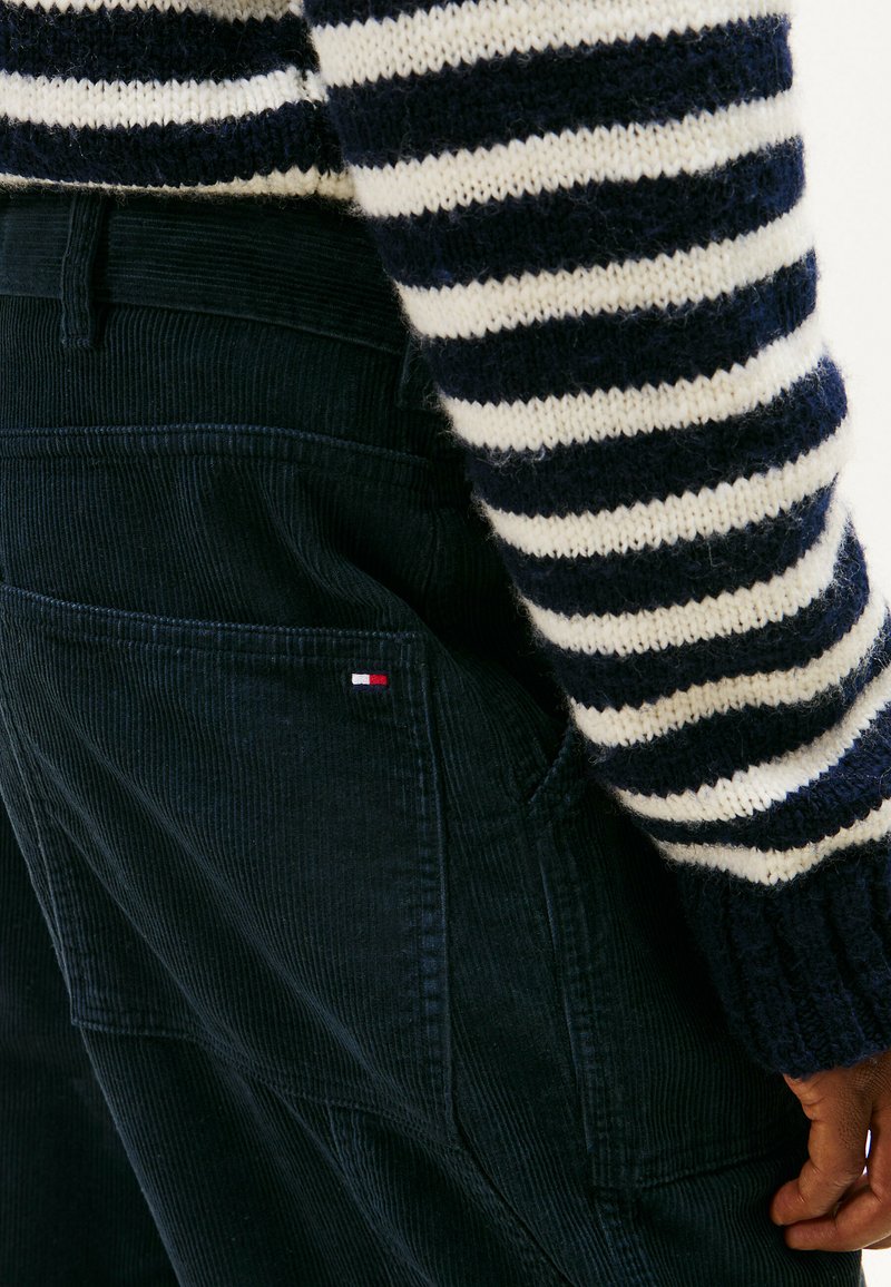 Navy corduroy overalls feature textured fabric, side pockets, and a small red, white, and blue logo. Striped knit sweater complements overall.