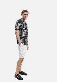 Camisa de manga corta con patrón en blanco y negro y diseños geométricos, combinada con pantalones cortos blancos y sandalias negras de tira.