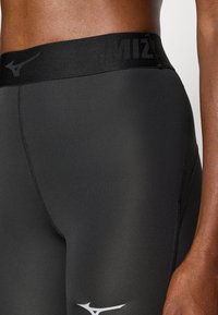 Sorte atletiske leggings har en glat tekstur, høj talje med "MIZUNO" logo og hvid logo detaljer nær nederste ben.