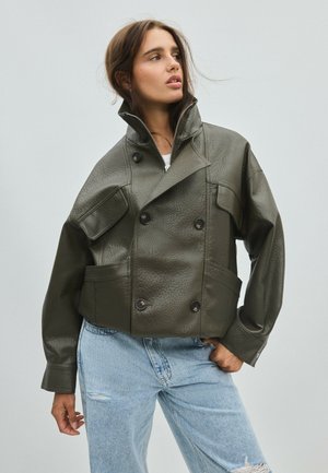 Femme en veste en cuir vert olive oversize avec de gros boutons et jean bleu clair déchiré, regardant sur le côté sur un fond uni.