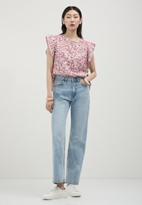 Donna che indossa un blusa senza maniche floreale rosa, jeans a vita alta azzurri chiari, sneakers bianche e braccialetti d'oro su uno sfondo chiaro e semplice.
