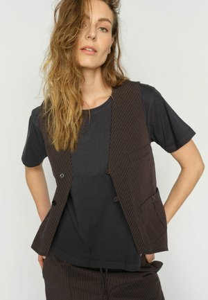 Frau mit welligem blondem Haar, die ein schwarzes T-Shirt sowie eine braune Nadelstreifenweste und passende Hose trägt, steht mit den Händen in den Taschen.