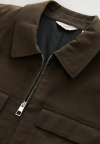 Chaqueta de lienzo marrón con cuello, cremallera frontal y dos bolsillos en el pecho. Tejido texturizado de un color sólido con forro en contraste.