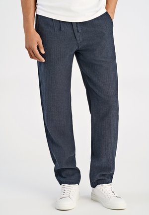Pantaloni blu scuro, strutturati, con vestibilità rilassata, vita elastica e polsini. Indossati con sneaker bianche e una t-shirt semplice.