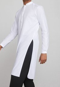 Kurta blanca de manga larga con cuello mao, tapeta con botones y aberturas laterales; combinada con pantalones oscuros para contraste. Textura de tela lisa.