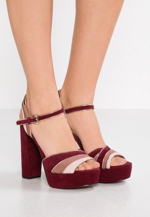 Bordowe aksamitne platformy na obcasie z szerokim paskiem i klamrą przy kostce. Charakteryzują się kolorową kompozycją w odcieniach bordowego i różowego z złotymi akcentami.