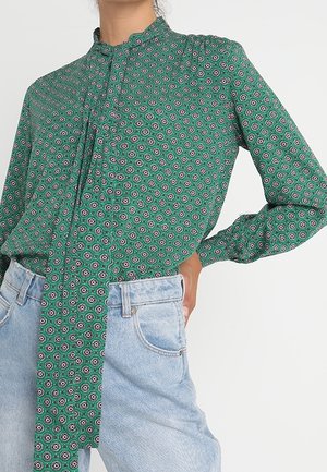 Grüne, gemusterte Langarmbluse mit Frontknoten, kombiniert mit hellblauen, hoch taillierten Jeans, die Hand ruht auf der Hüfte.