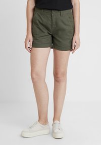 Green cotton shorts with a fitted design, rolled cuffs, front pockets, and a button closure. Paired with light-colored platform shoes.