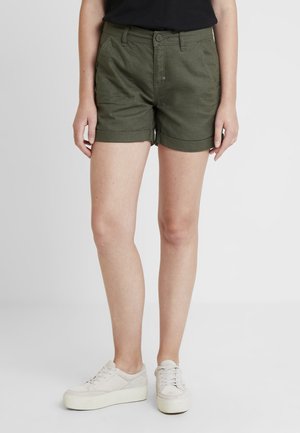 Shorts en coton verts avec une coupe ajustée, des poignets retroussés, des poches avant et une fermeture à bouton. Associés à des chaussures à plateforme de couleur claire.