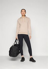 Sweatshirt bege com capuz e bolso frontal, leggings pretas e ténis desportivos pretos. A segurar uma grande bolsa de ginásio preta. Design simples e funcional.