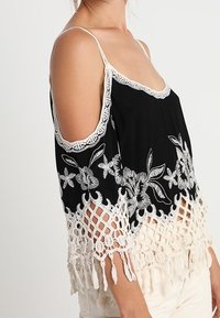 Haut noir épaules dénudées avec broderie florale blanche et détails en crochet, présentant une coupe ample et un ourlet à franges.