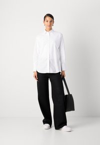 Camicia bianca con dettagli decorativi, abbinata a pantaloni neri a gamba larga con sottili decorazioni e sneakers bianche.