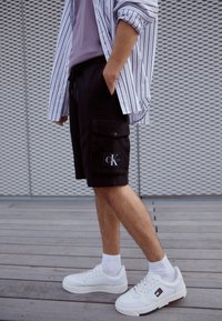 Černé cargo šortky s bočními kapsami, doplněné světle fialovou košilí a bílými teniskami. Tento outfit kombinuje kontrastní textury a barvy.