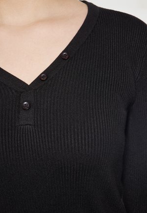 Schwarzes geripptes Strickoberteil mit V-Ausschnitt, ausgestattet mit zwei braunen Knöpfen am Kragen, betont die Textur und das taillierte Design.