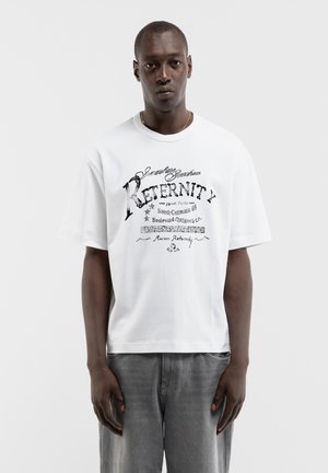 Mann trägt ein weißes T-Shirt mit schwarzem Aufdruck und graue Jeans vor einem einfarbigen weißen Hintergrund, steht und schaut nach vorne.