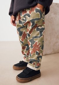 Pantalon camouflage vert, marron et beige, coupe ample avec revers roulés, associé à des baskets noires à scratchs et semelles en gomme.
