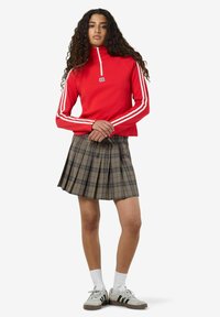 Felpa rossa con zip e strisce bianche sulle maniche, abbinata a una gonna a pieghe a scacchi. La modella indossa sneakers grigie e calze bianche.