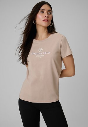 Kobieta o długich ciemnych włosach, nosząca kolczyki koła, beżowy t-shirt „Victory Club” oraz czarne spodnie, patrząca na bok na tle jednolitego tła.