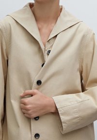 Manteau beige avec un grand col, doté de boutons noirs, à manches longues et à la texture douce, partiellement boutonné, tenu d'une main.