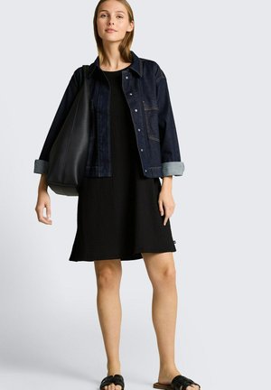 Frau trägt schwarzes Kleid und hochgekrempelte dunkle Jeansjacke, trägt große schwarze Tasche auf der Schulter, steht vor einfachem Hintergrund.