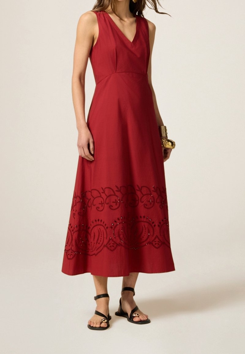 Robe maxi sans manches rouge avec un décolleté en V, dotée de broderies au niveau de l'ourlet et associée à des sandales plates noires.