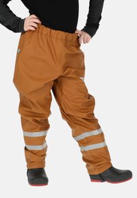 Waterdichte bruine broek met elastische tailleband, reflecterende strepen en een losse pasvorm, gecombineerd met zwarte rubberen laarzen met rode zolen.