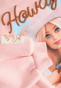 Tissu rose avec une illustration d'une poupée Barbie, yeux bleus et cheveux blonds, montrant un bras levé et le mot "Home" en lettres marron en relief.