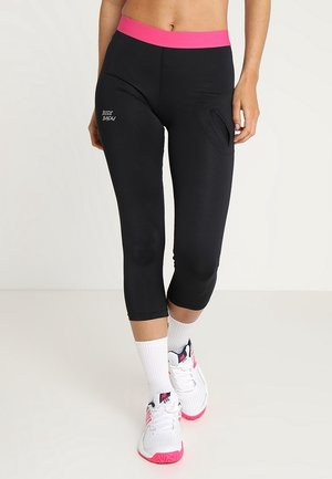 Femme portant des leggings noirs coupés avec une ceinture rose et des chaussures de sport blanches avec des touches roses, debout sur un sol clair.
