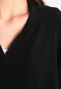Cardigan nero con scollatura a V, caratterizzato da una texture a coste e un tessuto morbido, che mostra parzialmente un top bianco sottostante.
