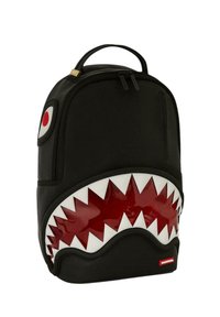 Mochila negra con diseño de boca de tiburón, que presenta dientes rojos brillantes y una superficie texturizada. Incluye un asa superior y cierres de cremallera.