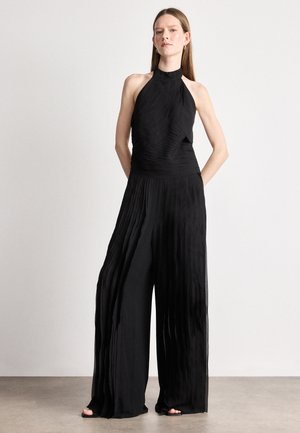 Combinaison noire à encolure halter avec texture plissée, jambes larges et corsage ajusté. Caractéristiques d'un col montant et tissu fluide avec peu d'accents en métal.