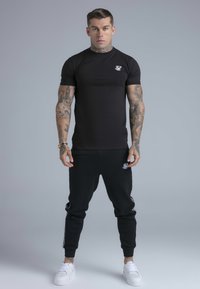 Musta istuva t-paita, jossa on lyhyet hihat ja pieni logo, yhdistettynä mustiin vapaa-ajan housuihin, joissa on sivuilla logot, sekä valkoiset urheilukengät.