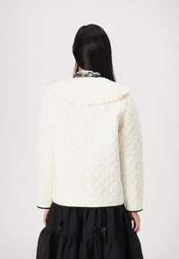 Καπιτοναρισμένο ιβουάρ σακάκι με ρυτιδωτό γιακά, μακριά μανίκια και αντίθετη μαύρη μπορντούρα, συνδυασμένο με μίντι μαύρη φούστα με επίπεδες ριπές και φιόγκους.