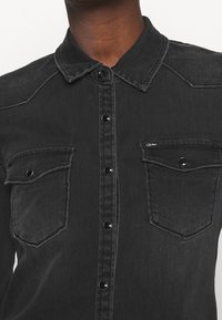 Primo piano di una persona che indossa una camicia di denim nera con bottoni automatici neri e due tasche a pattina anteriori.