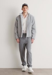 Chaqueta de cremallera gris claro con un forro suave, combinada con pantalones de chándal grises y zapatillas blancas. El modelo lleva una gorra gris a juego.
